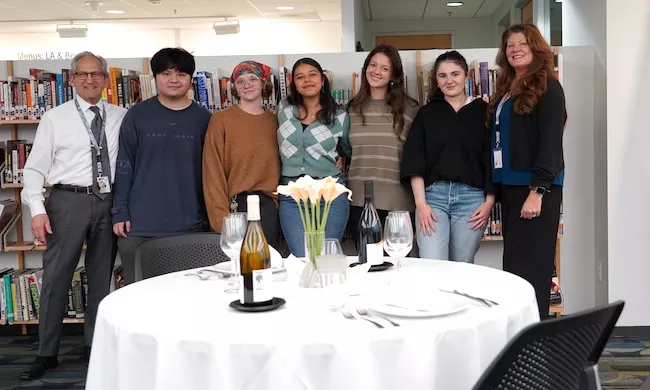 ICE Culinary Management students smile in a library behind a set table with Instructor Paul Sherman, a man in a white shirt and grey pants and Restaurant & Culinary Management Director Mishel LeDoux, a woman with brown hair in a black blazer and black pants