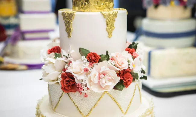 A wedding cake with pink and red flowers and gold leaf decorated during a class in culinary school.