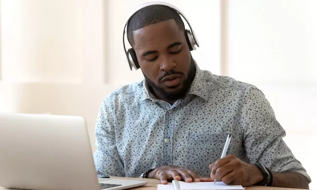 A male student wearing heaphones and a blue button down shirt takes notes on a notepad while in front of a laptop