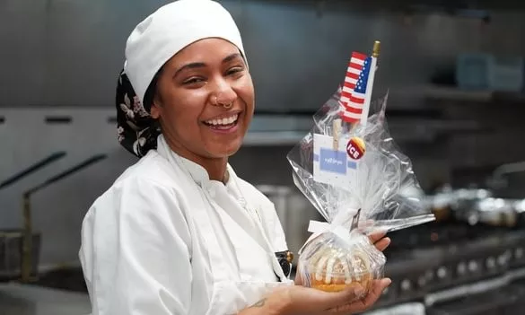 an ICE student and US veteran in a chef jacket and hat holds up a wrapped baked good with an american flag and an ICE logo in a professional kitchen