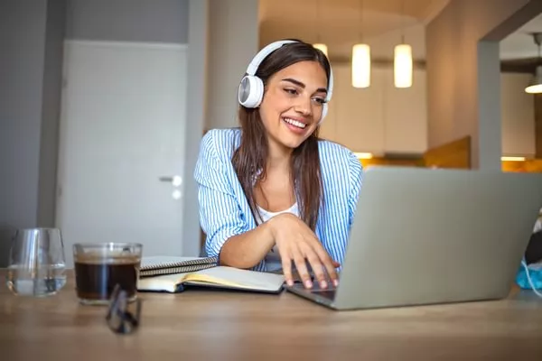 A female student wearing heaphones and a blue shirt takes notes on a notepad while in front of a laptop