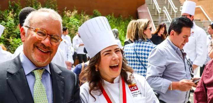 ICE graduate Alessandra Ciuffo, a brunette woman, smiles in an ICE white chef's coat and hat next to Rick Smilow, a man in a blue suit wearing glasses