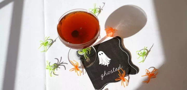 A Black Manhattan cocktail, a dark brown drink with a cherry at the bottom, sits in a coupe glass on a black napkin with a cartoon ghost on white table with a green and orange plastic spider rings surrounding it