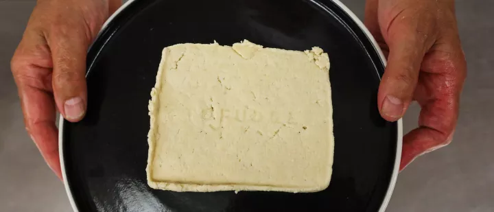 White block of tofu on a dark plate, hands on either side holding plate