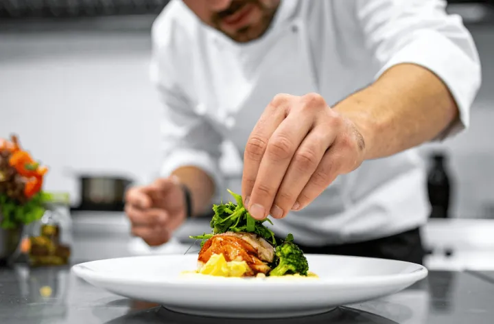 A close-up shot of a chef in a white uniform carefully plating a dish in a kitchen. The chef uses their fingers to precisely place fresh green microgreens on top of a beautifully presented meal, which includes meat, broccoli, and a purée on a wide white plate.