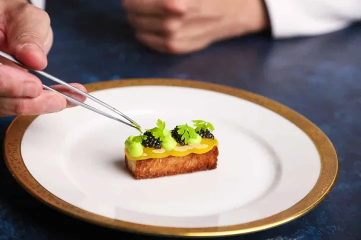 a chef uses large tweezers to place an herb leaf on a dish with a stack of bread, lemon gel and blackberries
