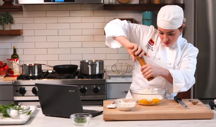 A Culinary Arts & Food Operations associate degree student grinds pepper into a glass bowl with flour and eggs while looking at a laptop