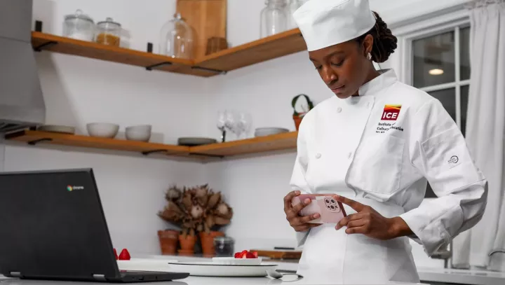 Culinary student in white chef coat and hat stands in home kitchen and photographs dish in front of laptop