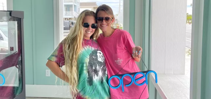 Anna Beth and Lauren stand together smiling and holding an "open" sign